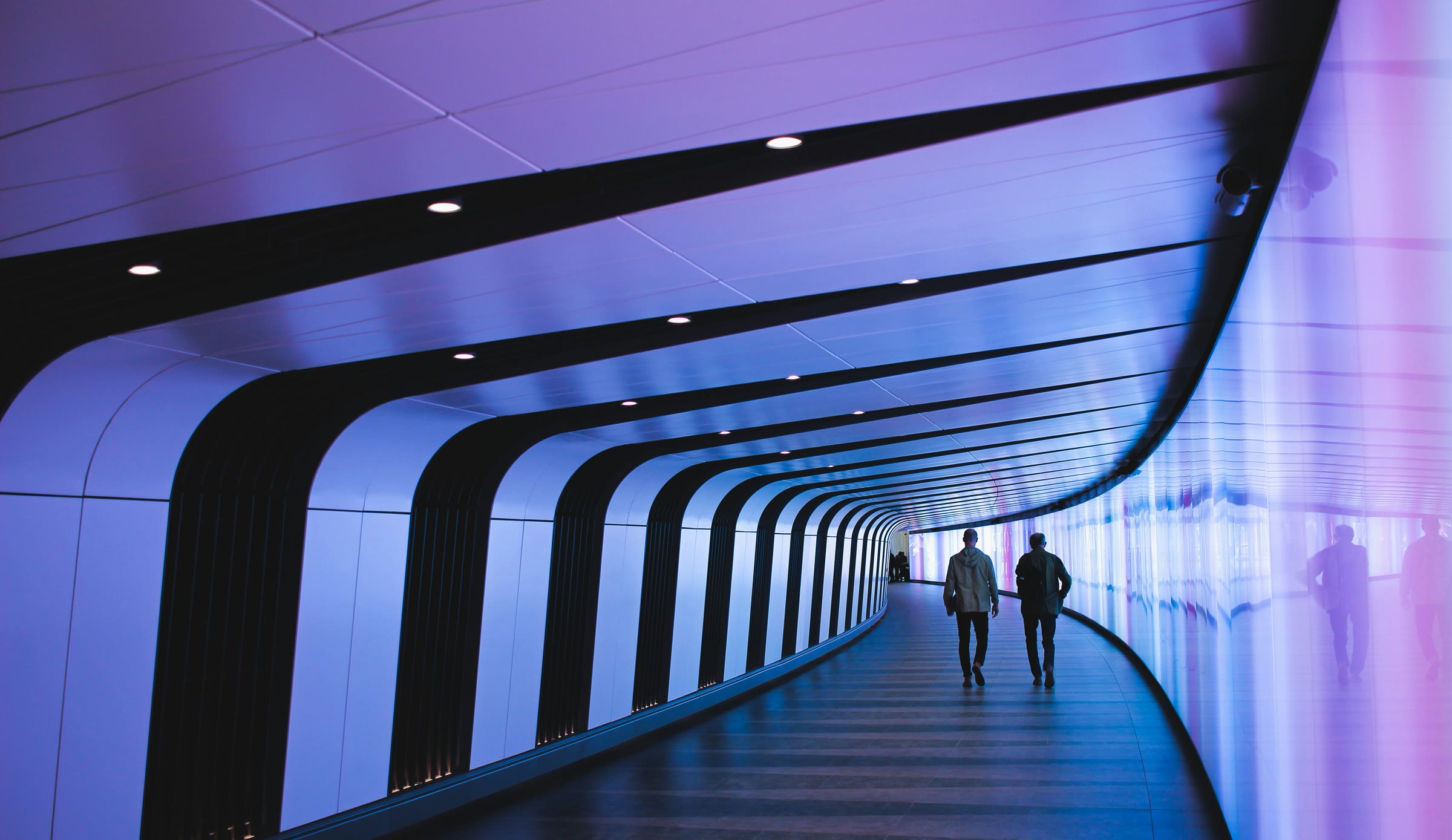 Two people walking away down a curved corridor washed in violet and blue light